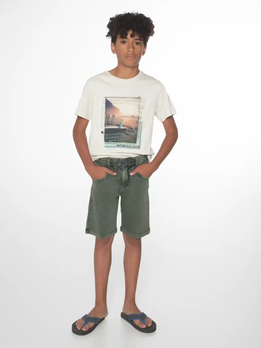 Un jeune homme aux cheveux bouclés se tient debout devant un fond blanc. Il porte un t-shirt avec un motif de plage, un short vert et des tongs. Il a les mains dans les poches de son short.