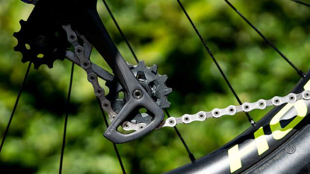 Nahaufnahme einer Fahrradkette und des Umwerfers vor grünem Hintergrund. Die Kette ist silbern und sauber.