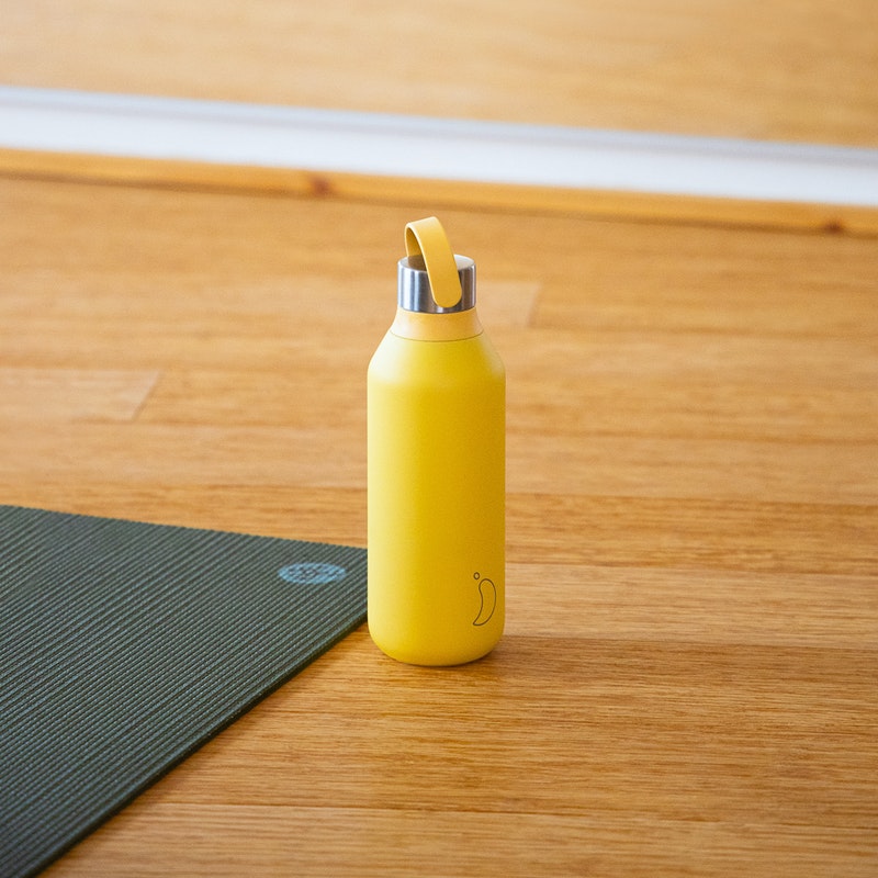 Gelbe Chilly's Series 2 Flasche (1000ml) steht auf einem Holzboden neben einer grünen Yogamatte.