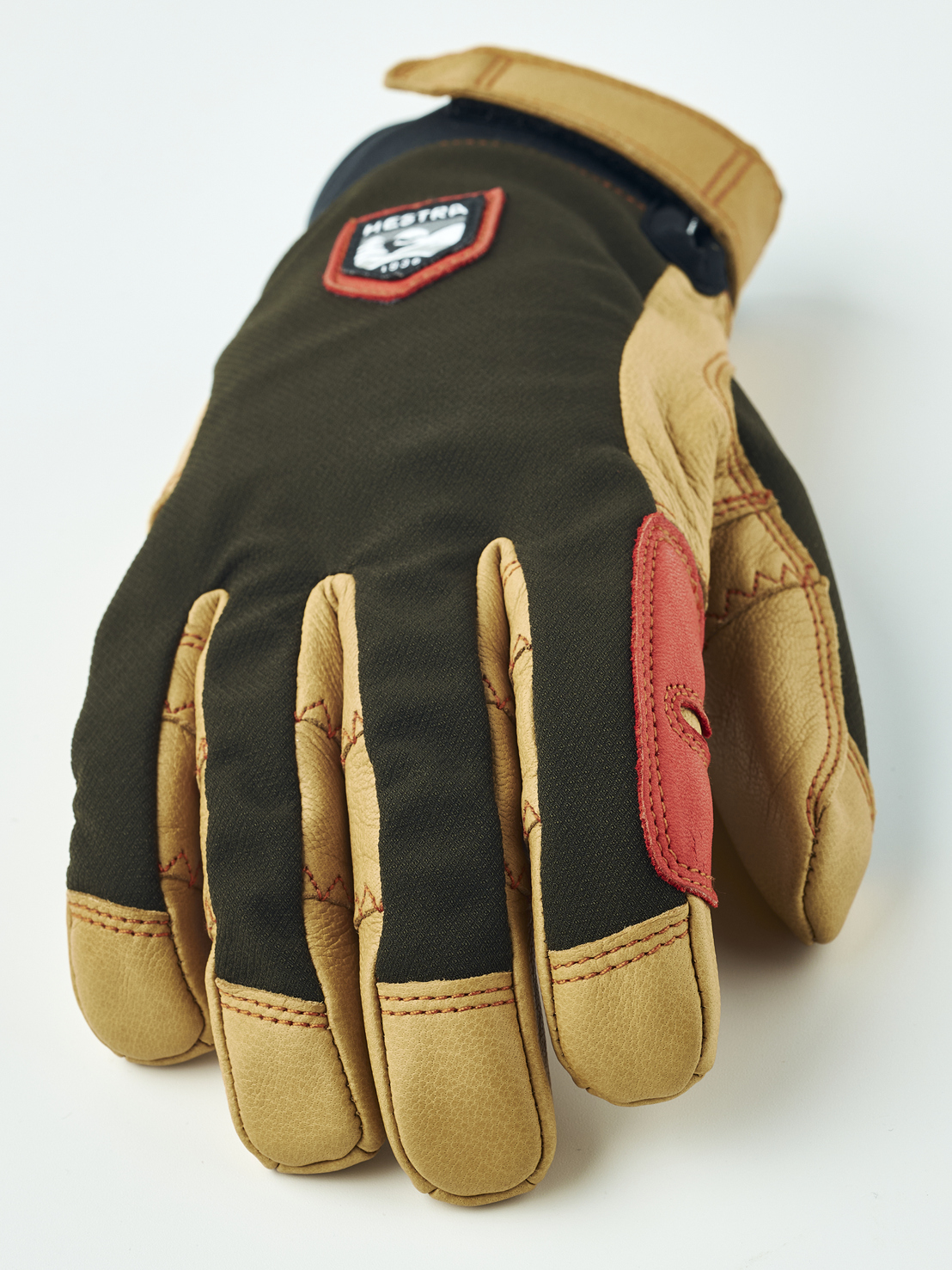 Nahaufnahme eines braunen Hestra-Handschuhs mit beigen Lederdetails und rotem Patch. Das Markenlogo ist auf dem Handrücken zu sehen.