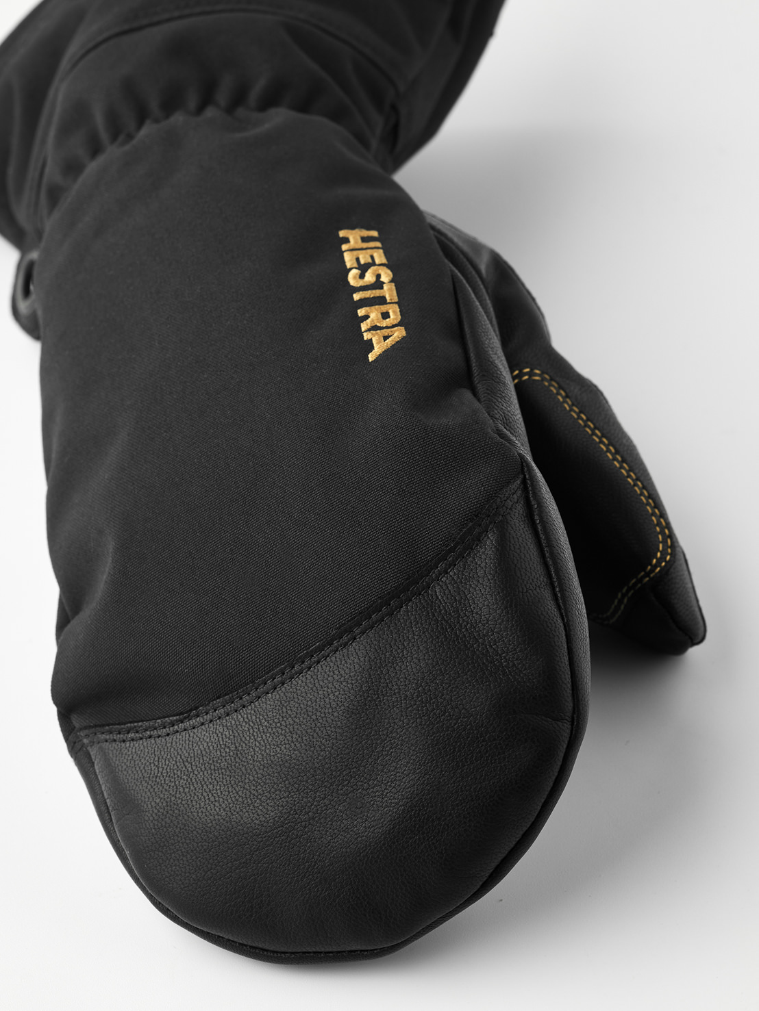 Hestra Moufle noire Vue détaillée d'une moufle noire Hestra en cuir et tissu, avec le logo doré Hestra sur le dos de la main.
