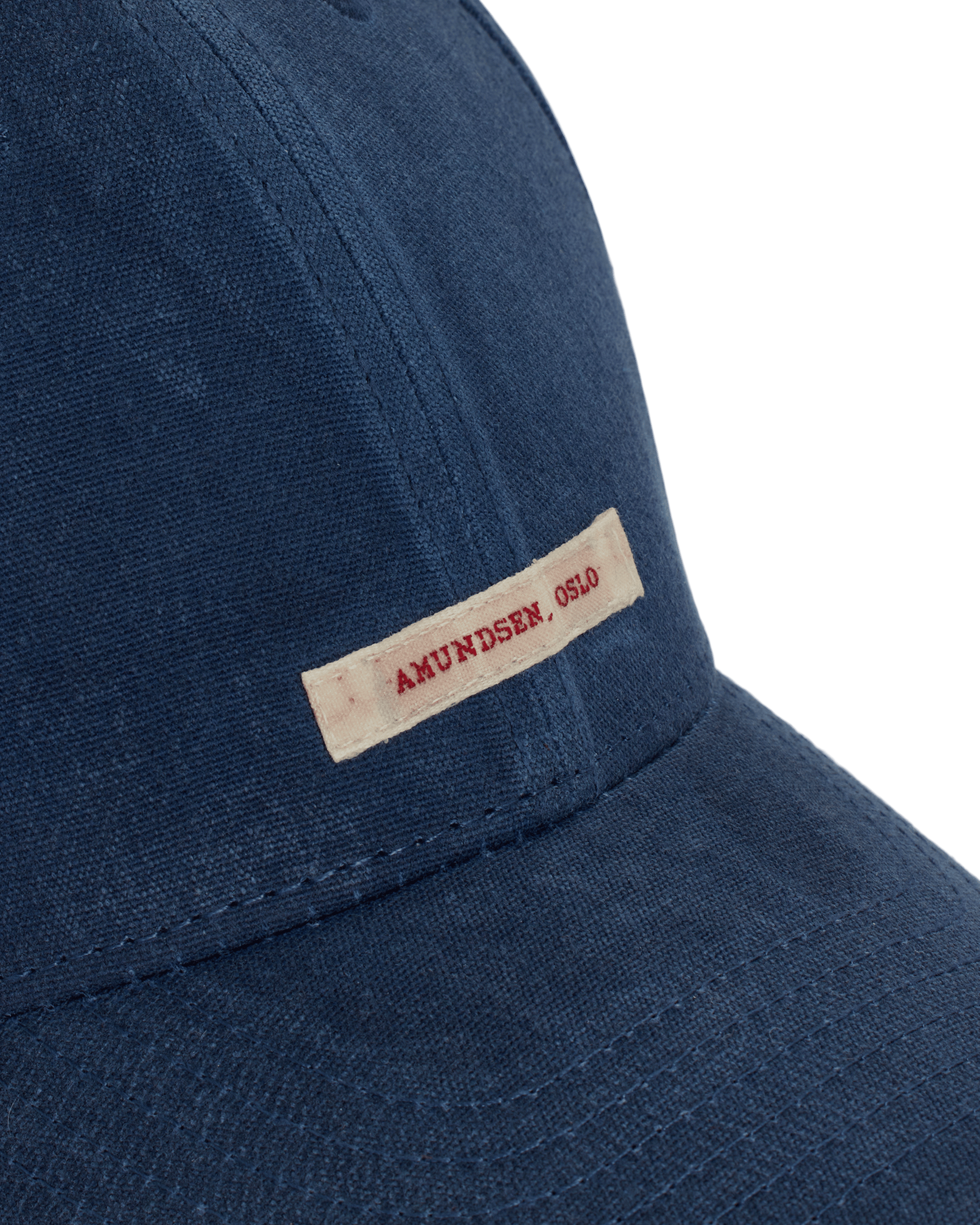 Eine blaue Baseballkappe ist aus einem groben Stoff gefertigt. Ein rechteckiges cremefarbenes Etikett ist an der Seite der Kappe befestigt, mit dem Text „AMUNDSEN, OSLO“ in roter Schrift. Nähte akzentuieren den Schirm und die Seiten der Kappe.