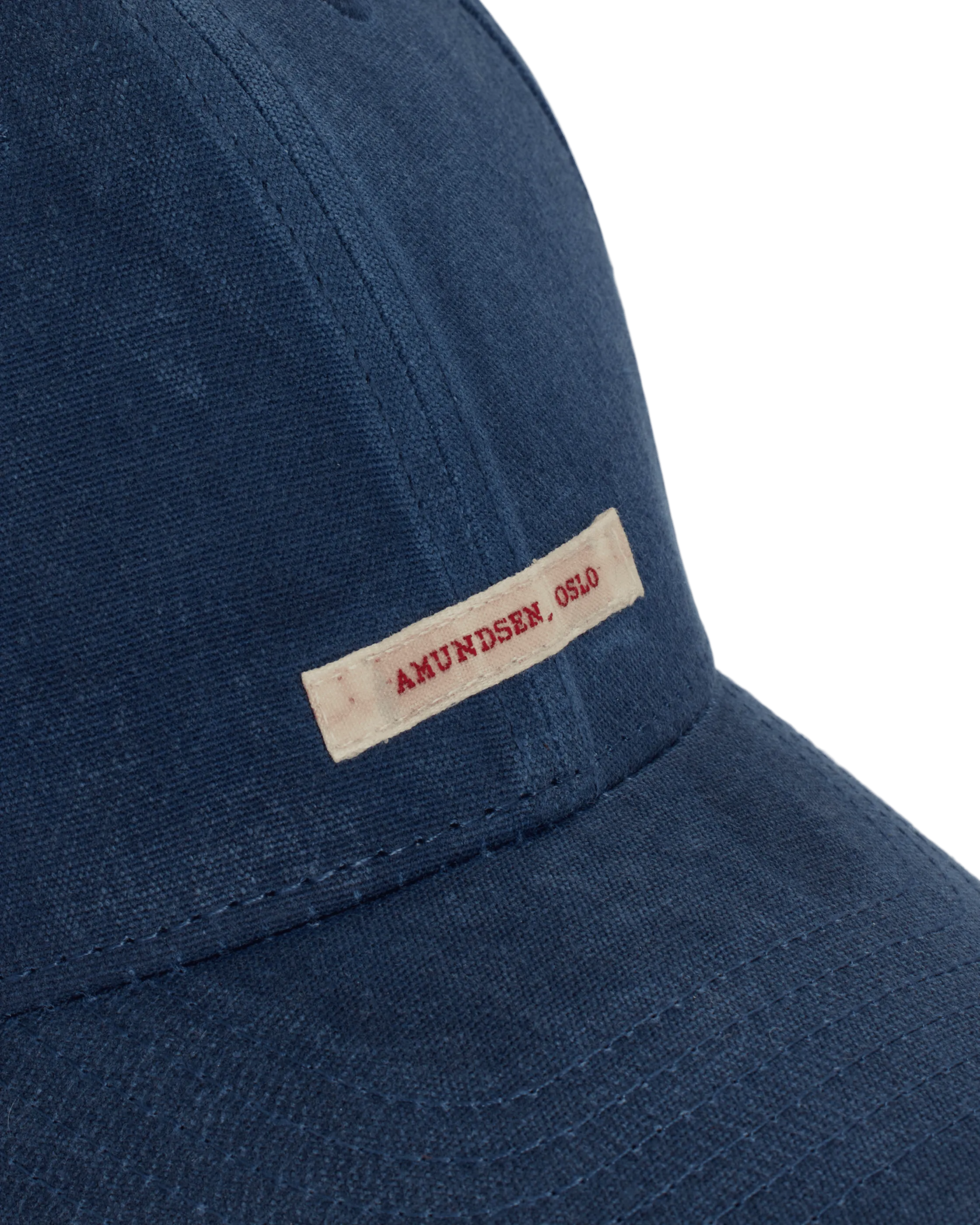 Un cappellino da baseball blu è realizzato in tessuto grezzo. Sul lato del cappellino è applicata un'etichetta rettangolare color crema con la scritta "Amundsen, OSLO" in rosso. Le cuciture accentuano la visiera e i lati del cappellino.