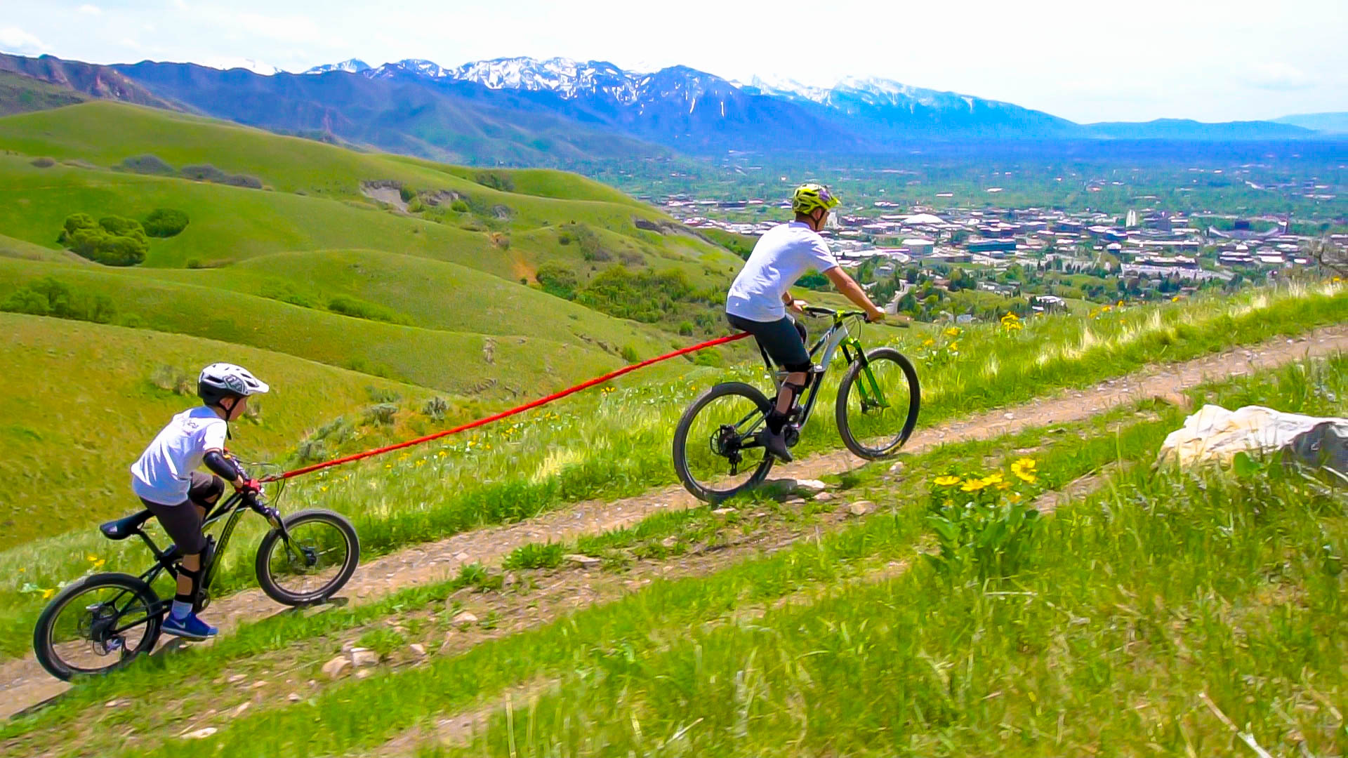 Zwei Kinder fahren mit dem Fahrrad bergauf, verbunden durch ein rotes Tow-Whee Abschleppseil vor einer grünen Hügellandschaft und Bergen.