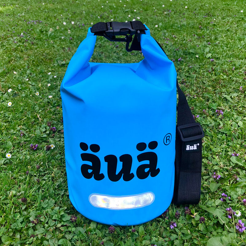 Blaue Dry Bag mit Logo auf grünem Gras, Tragegurt und reflektierendes Detail.
