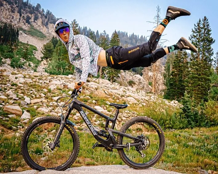 Uomo in equilibrio sulla mountain bike davanti alle montagne. Indossa una giacca con cappuccio, pantaloncini e calzini con un'atmosfera da "afterparty".