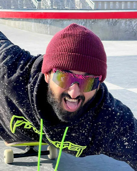 Uomo con berretto e occhiali fotocromatici, sdraiato sullo skateboard e con la lingua fuori.