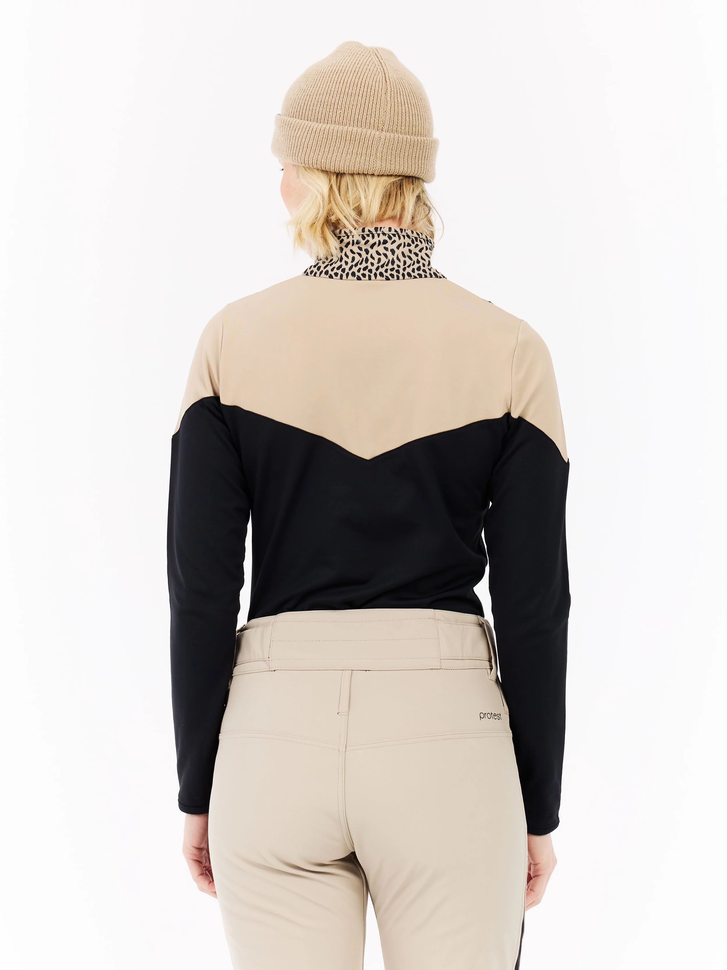 View from behind of a woman wearing a beige cap, a black and beige jacket and beige pants against a white background.