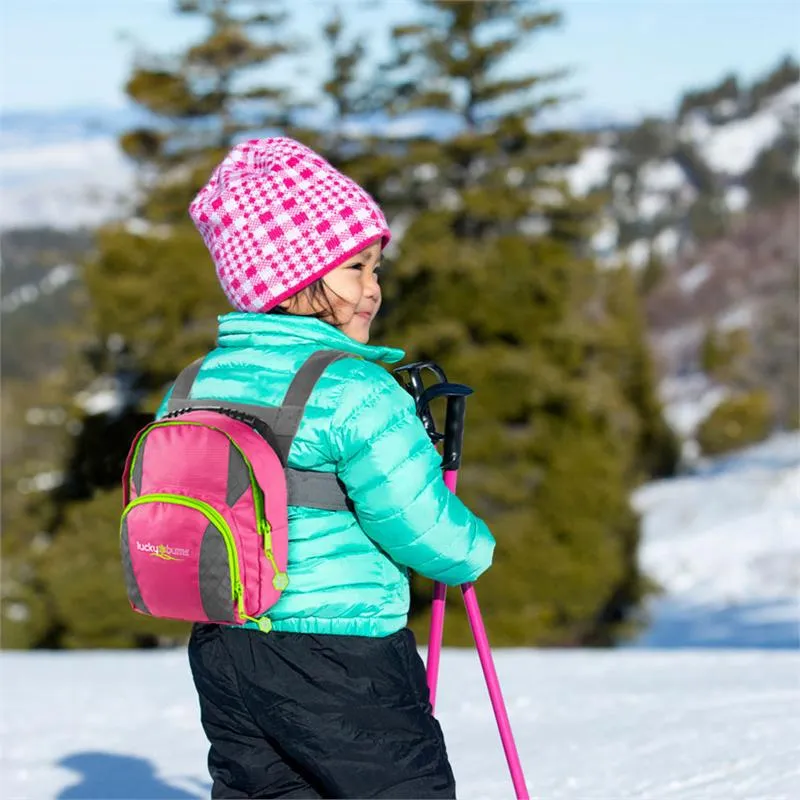 Kind mit Skitrainer, rosa Mütze, türkis Jacke, pink Rucksack und Skistöcken im Schnee.