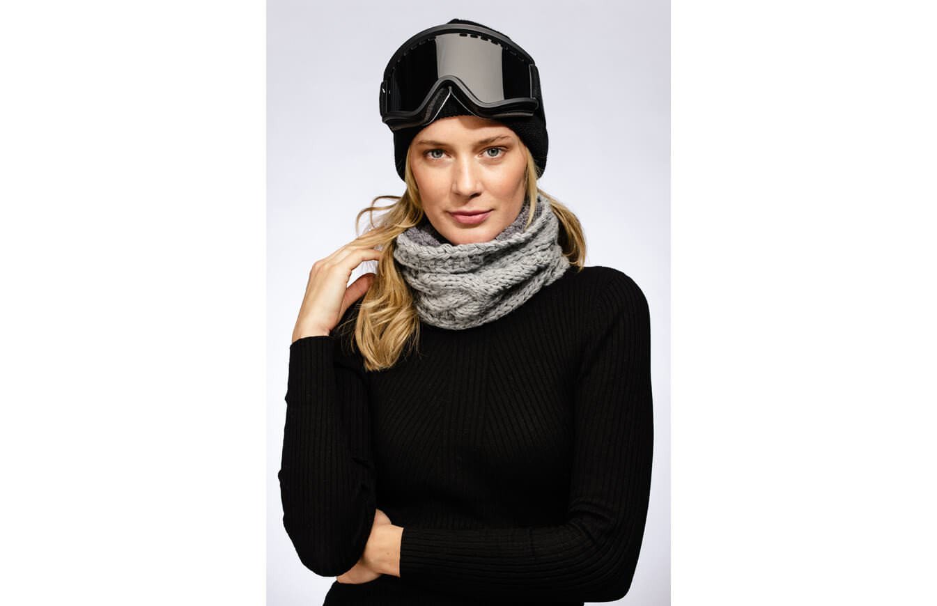Frau mit Skibrille und grauem Stroller Neckwarmer vor neutralem Hintergrund.