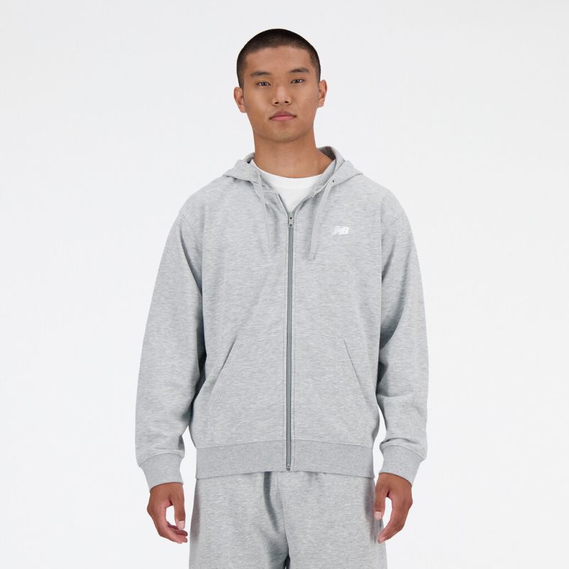 Grauer New Balance Hoodie Mann trägt einen grauen New Balance French Terry FZ Hoodie mit durchgehendem Reißverschluss und Kapuze. Vorne ist ein NB-Logo zu sehen.