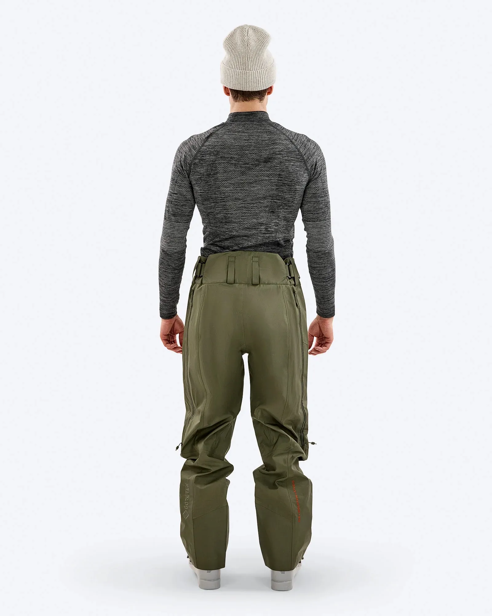 Un uomo di spalle, con un berretto grigio, una camicia grigia a maniche lunghe e pantaloni verde oliva. I pantaloni hanno delle cerniere ai lati.