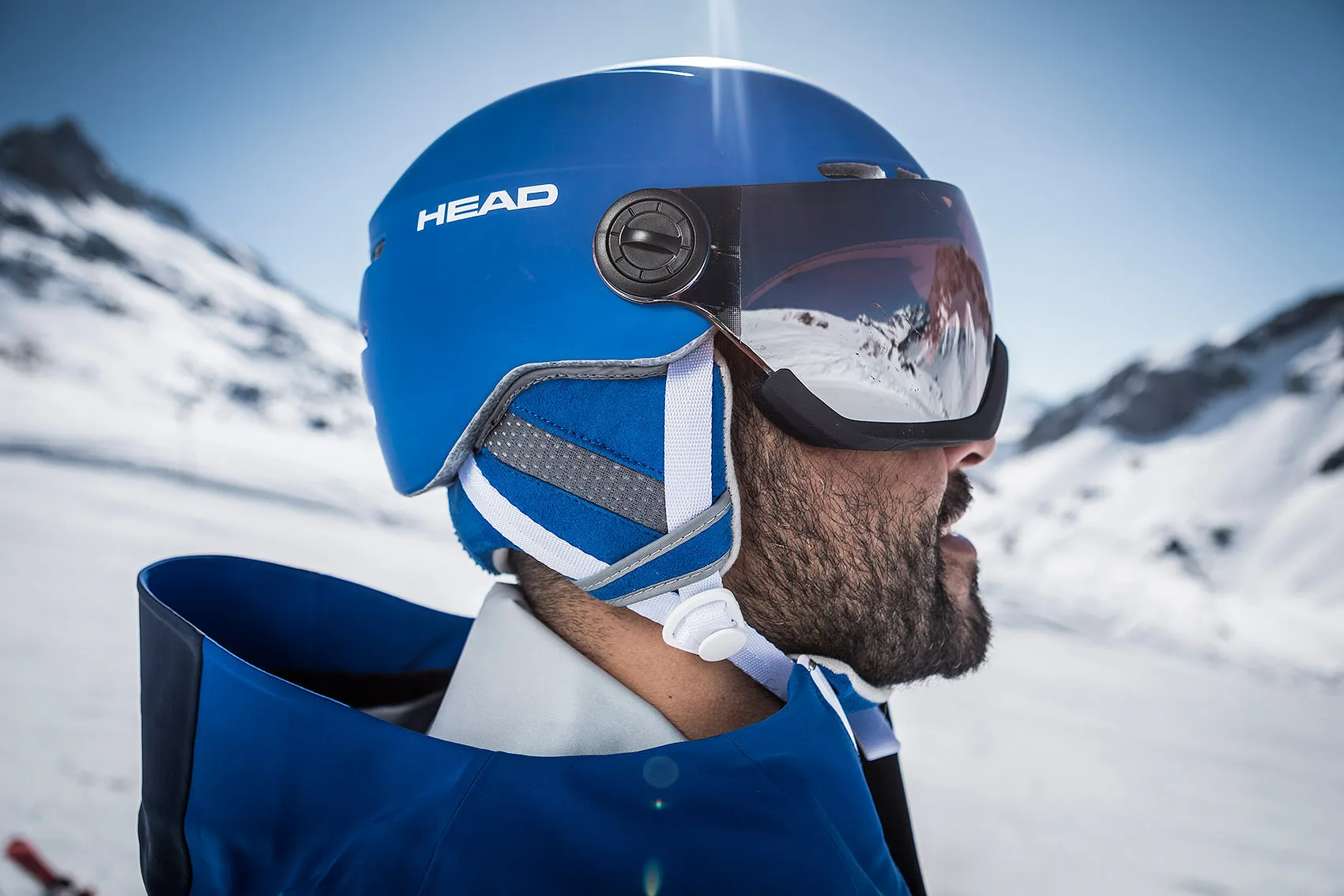 Vista di profilo di un uomo con casco da sci e visiera blu davanti a uno sfondo montano innevato.