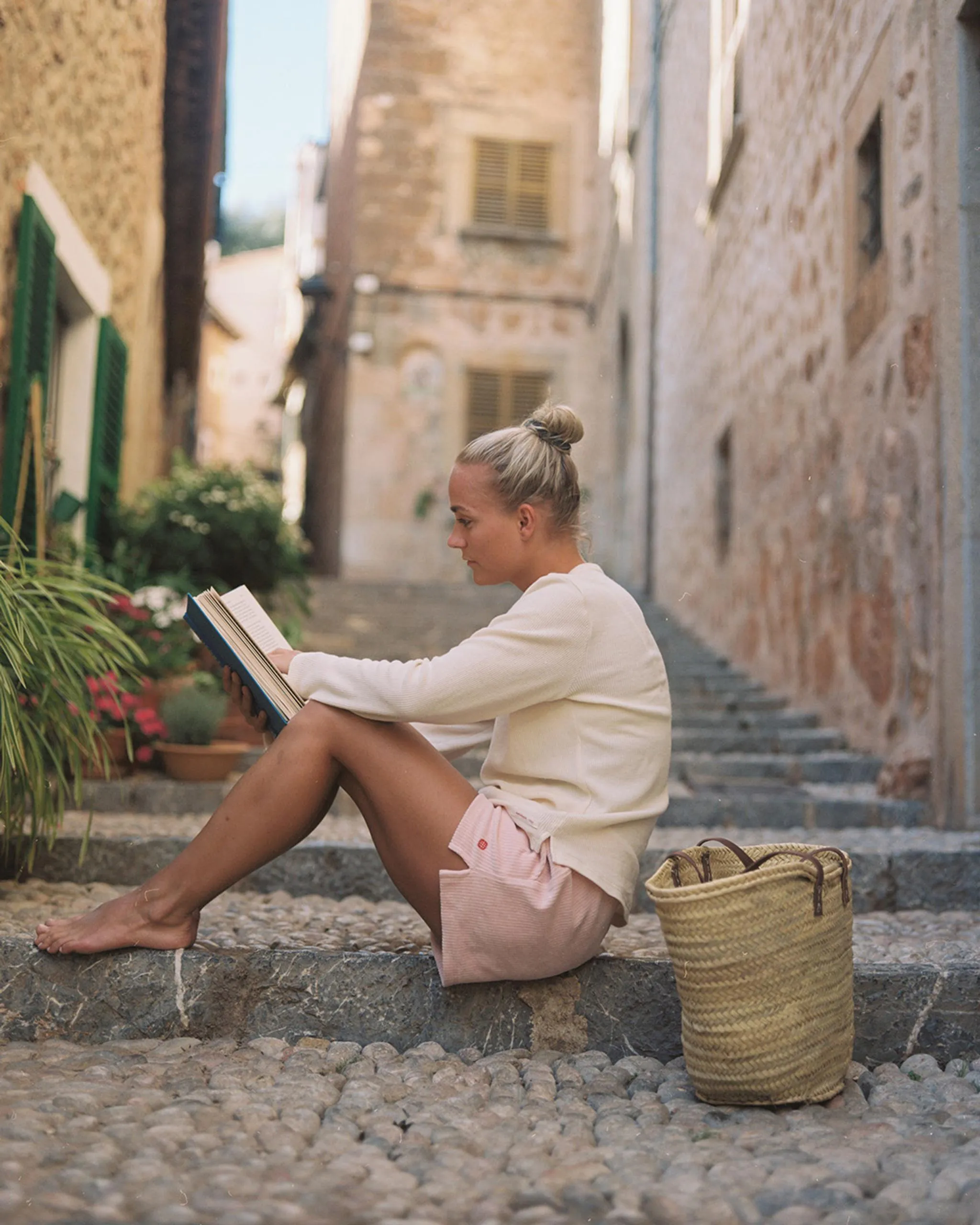 Eine junge Frau sitzt barfuß auf Steinstufen in einer Gasse und liest ein Buch. Neben ihr steht eine geflochtene Tasche.