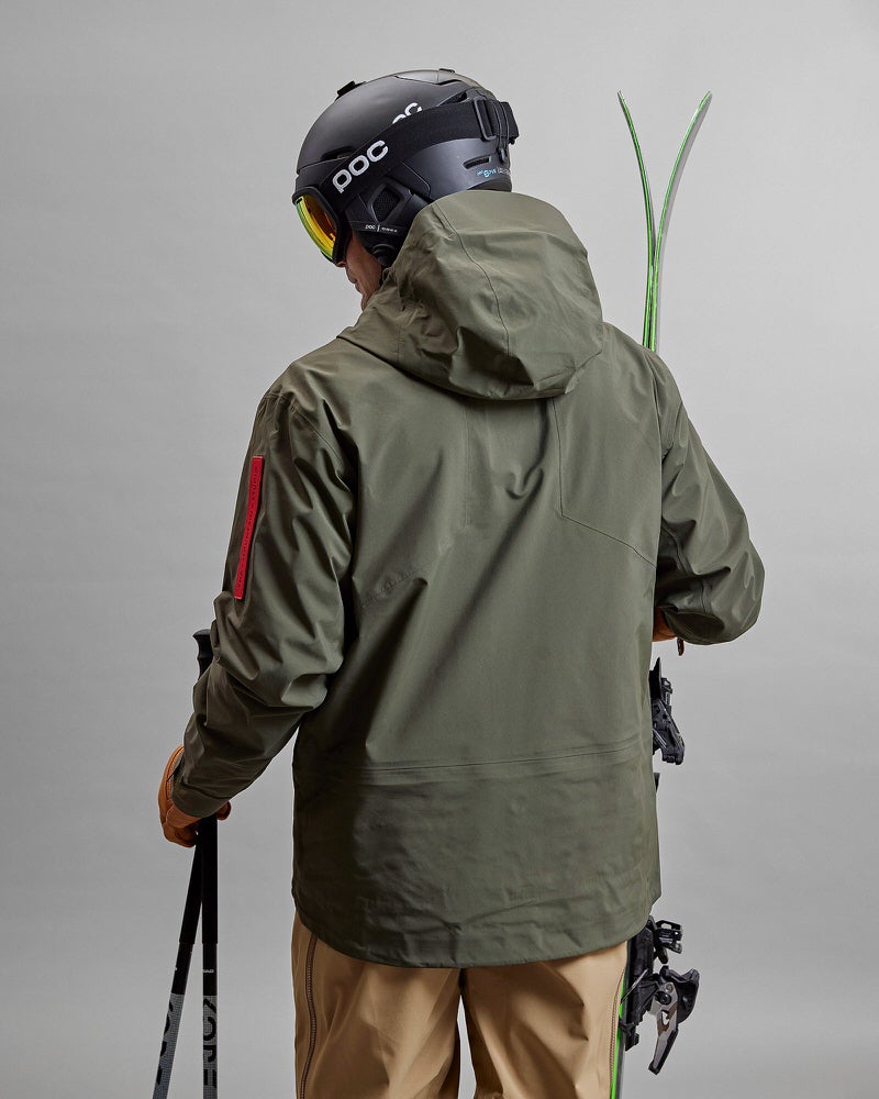 Skifahrer in GORE-TEX Jacke Person von hinten, in grüner Z-2 HD GORE-TEX Pro 3L Shell Jacke, Helm, Skier und Stöcke.