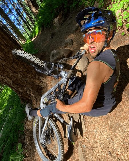 Mountain biker with helmet and amber-colored glasses sits on a forest path next to his bike. He looks into the camera.