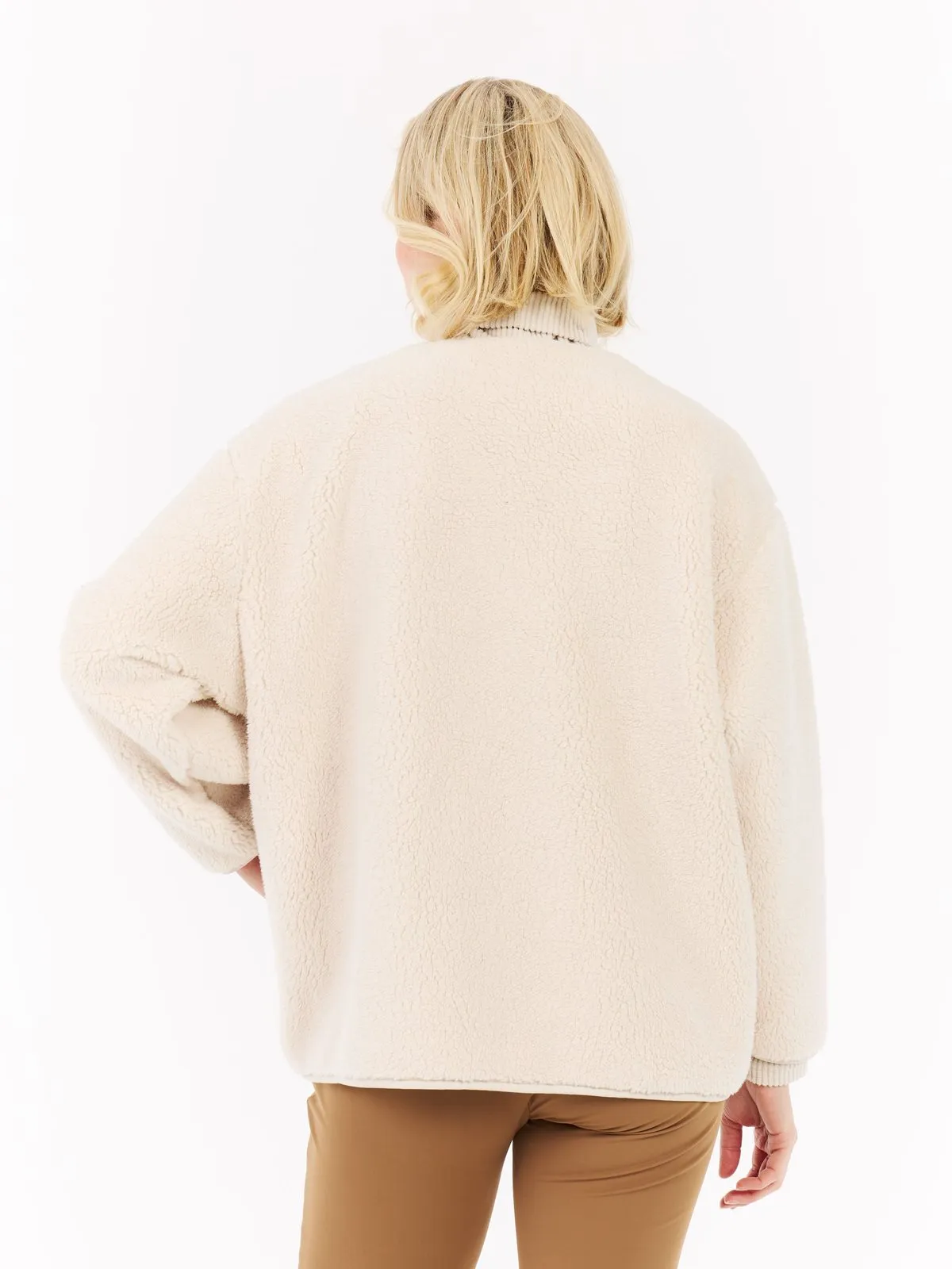 Vista posteriore di una donna con capelli biondi che indossa una giacca di pelliccia di orsacchiotto color crema e pantaloni marroni su sfondo bianco.