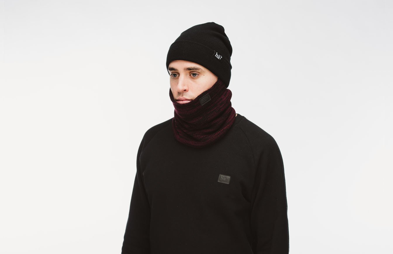 Ein Mann mit schwarzer Mütze und Melange Bordeaux Neckwarmer vor neutralem Hintergrund.