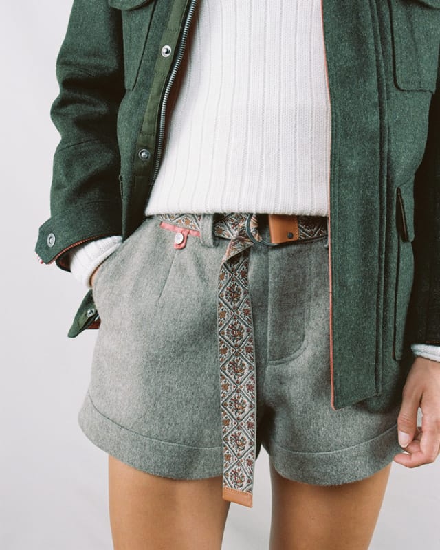 Eine Person trägt eine grüne Jacke, einen weißen Pullover und graue Shorts mit einem gemusterten Gürtel.