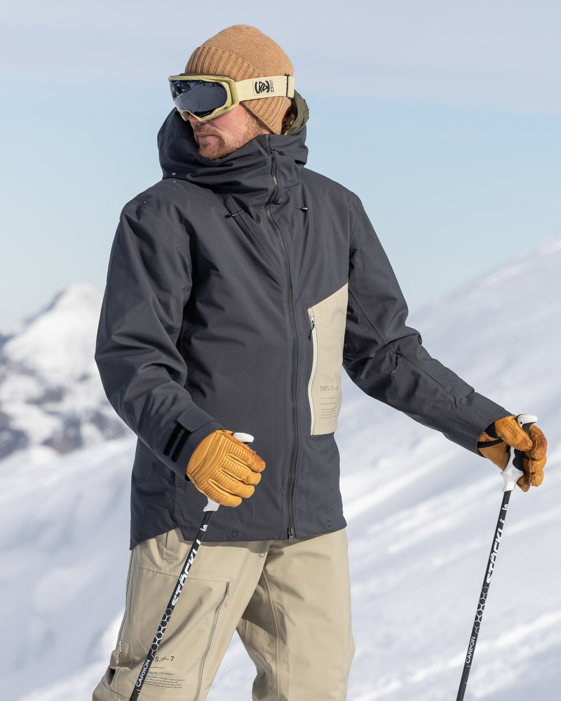 Mann mit Skibrille und Mütze trägt graue Ride Insulated Jacket und Handschuhe, hält Skistöcke im Schnee.