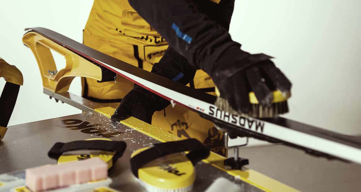 Eine Person bürstet einen Ski, der in einer gelben Vorrichtung befestigt ist. Wachsspäne liegen auf dem Tisch.
