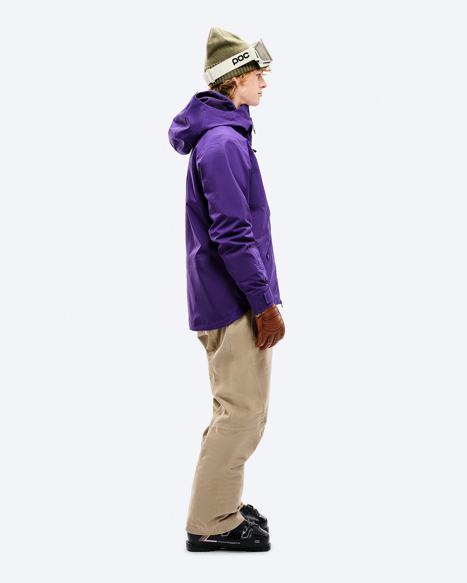 Veste GORE-TEX violette Personne vue de côté en veste Z-2 GORE-TEX violette, pantalon beige, chaussures de ski, bonnet et lunettes sur fond blanc.