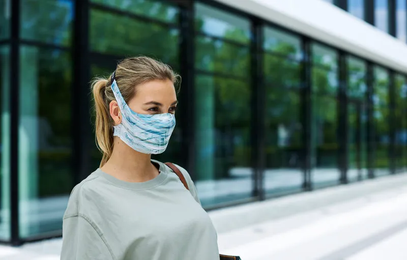 La femme porte un masque à filtre bleu clair Buff, fixé derrière la tête et la nuque par des rubans.