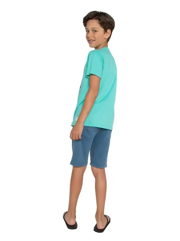 A boy with dark hair and braces looks over his shoulder at the camera. He is wearing a light blue T-shirt, blue shorts, and black flip-flops. The background is plain white.