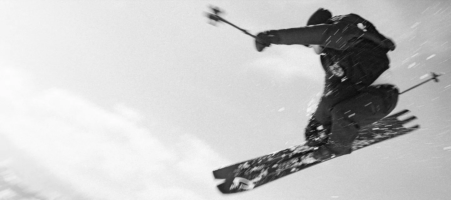 Photographie en noir et blanc d'un skieur en l'air, bras écartés et skis aux pieds, sur un fond clair.