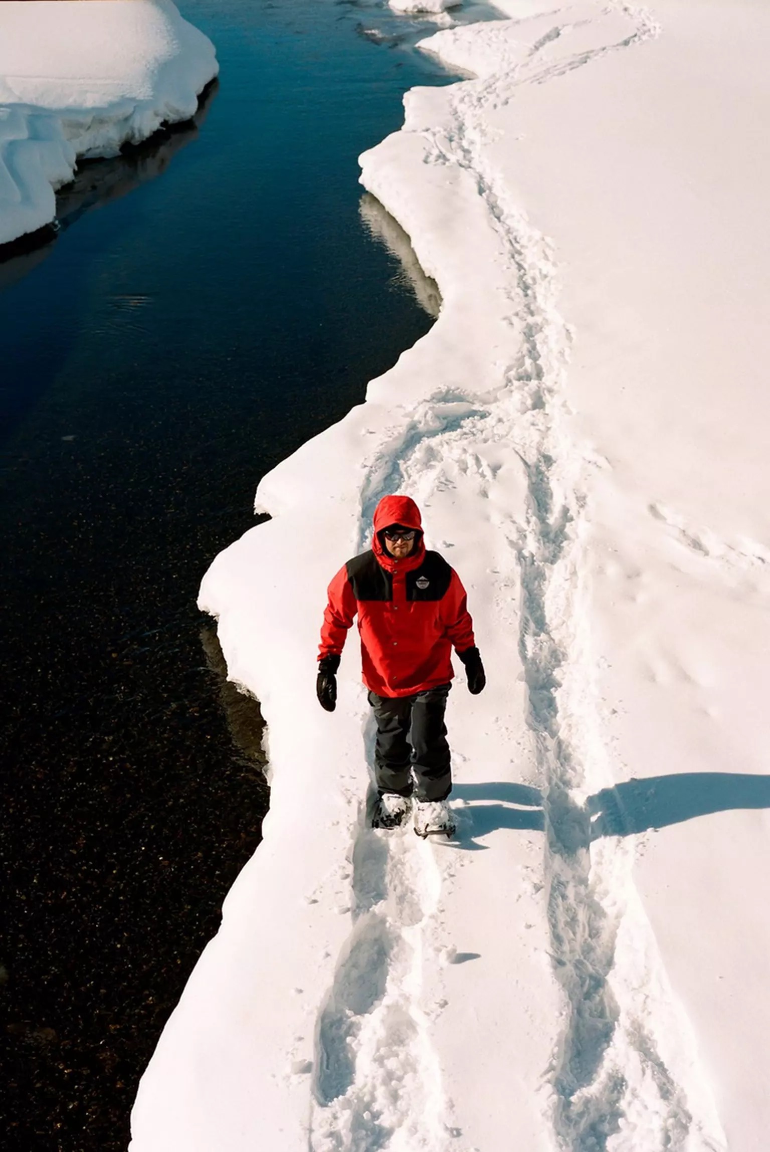 Homme marchant dans la neige Un homme portant des raquettes Tubbs Flex Alp marche sur un sentier enneigé près d'une rivière. Veste rouge.