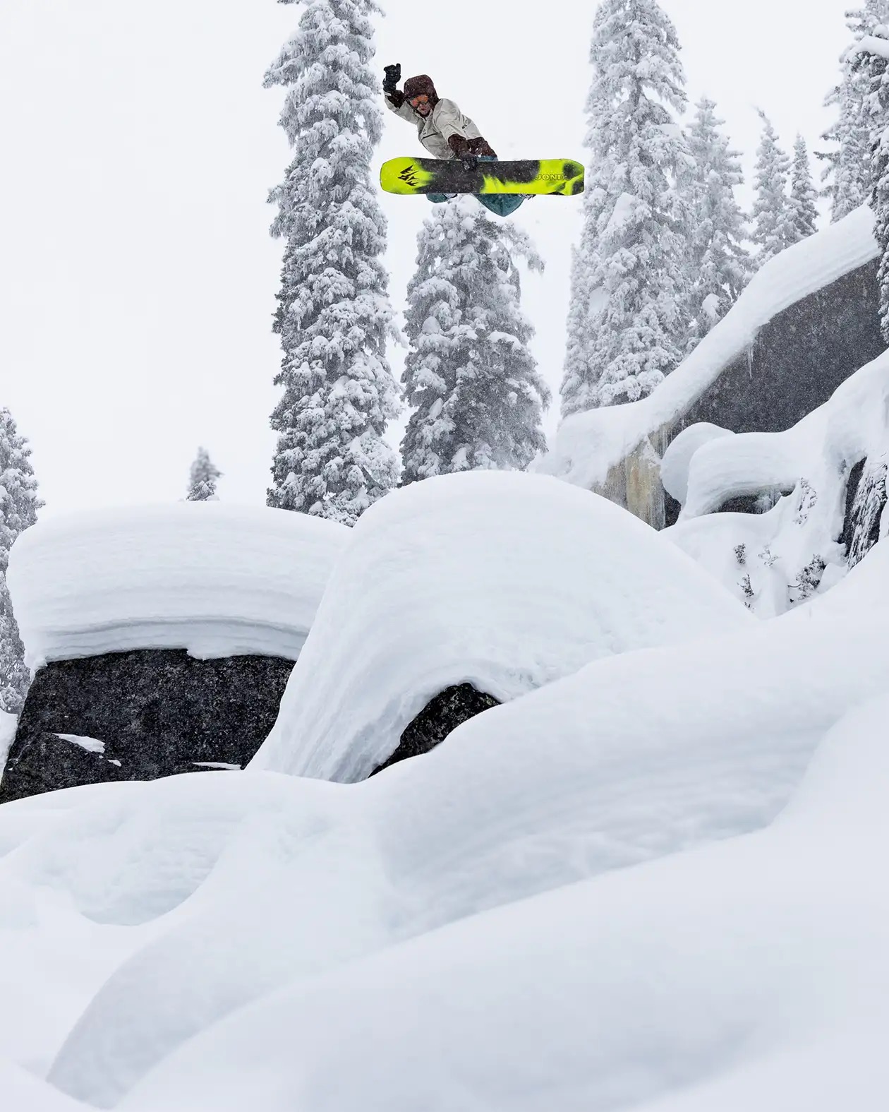 Snowboarder in der Luft vor verschneiten Bäumen und Felsen. Person mit Helm und Handschuhen auf gelb-schwarzem Board.
