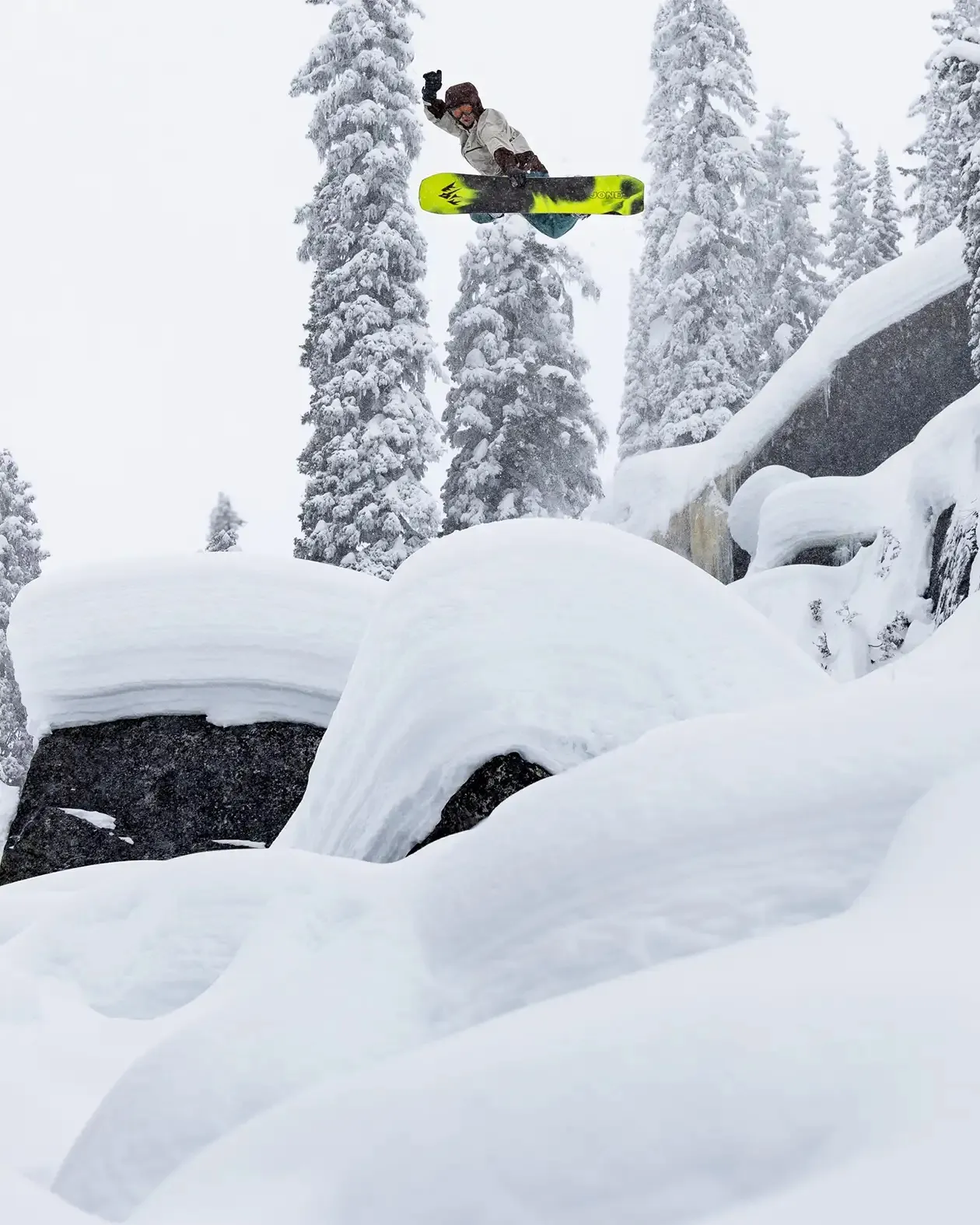Snowboarder in volo davanti ad alberi e rocce innevate. Persona con casco e guanti su una tavola gialla e nera.