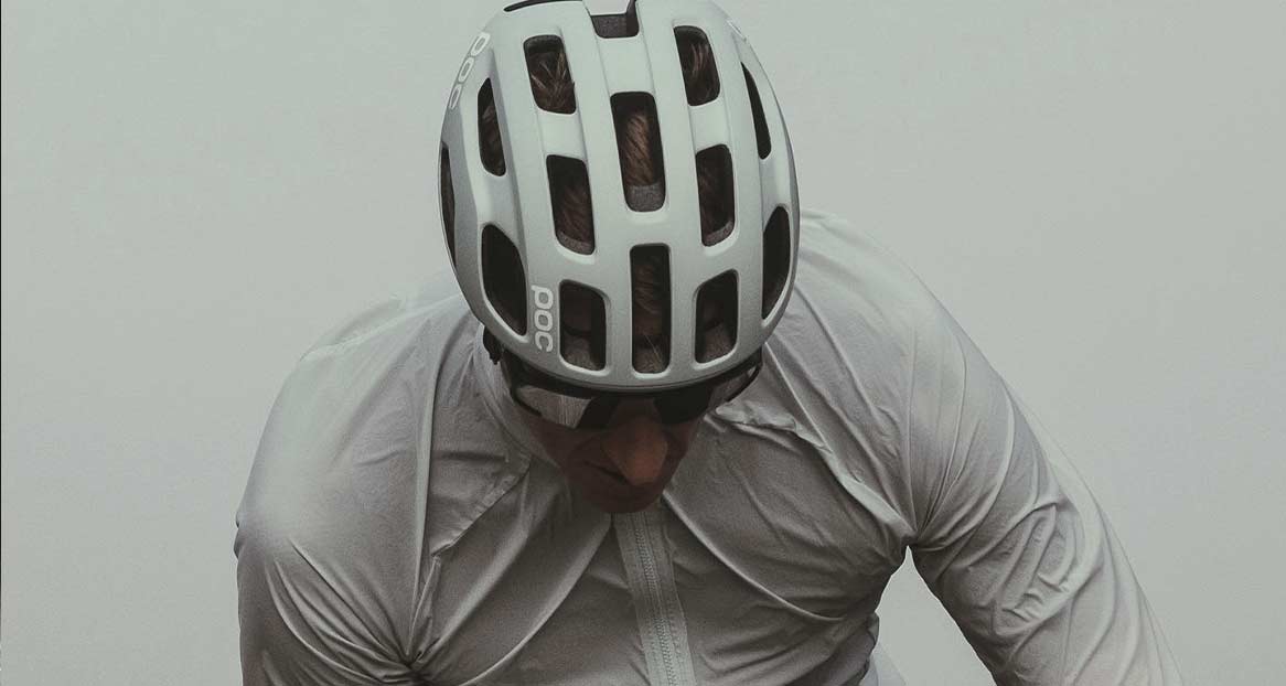 Un homme portant des lunettes de soleil et un casque de vélo blanc de la marque POC regarde vers le bas. Il porte une veste claire.