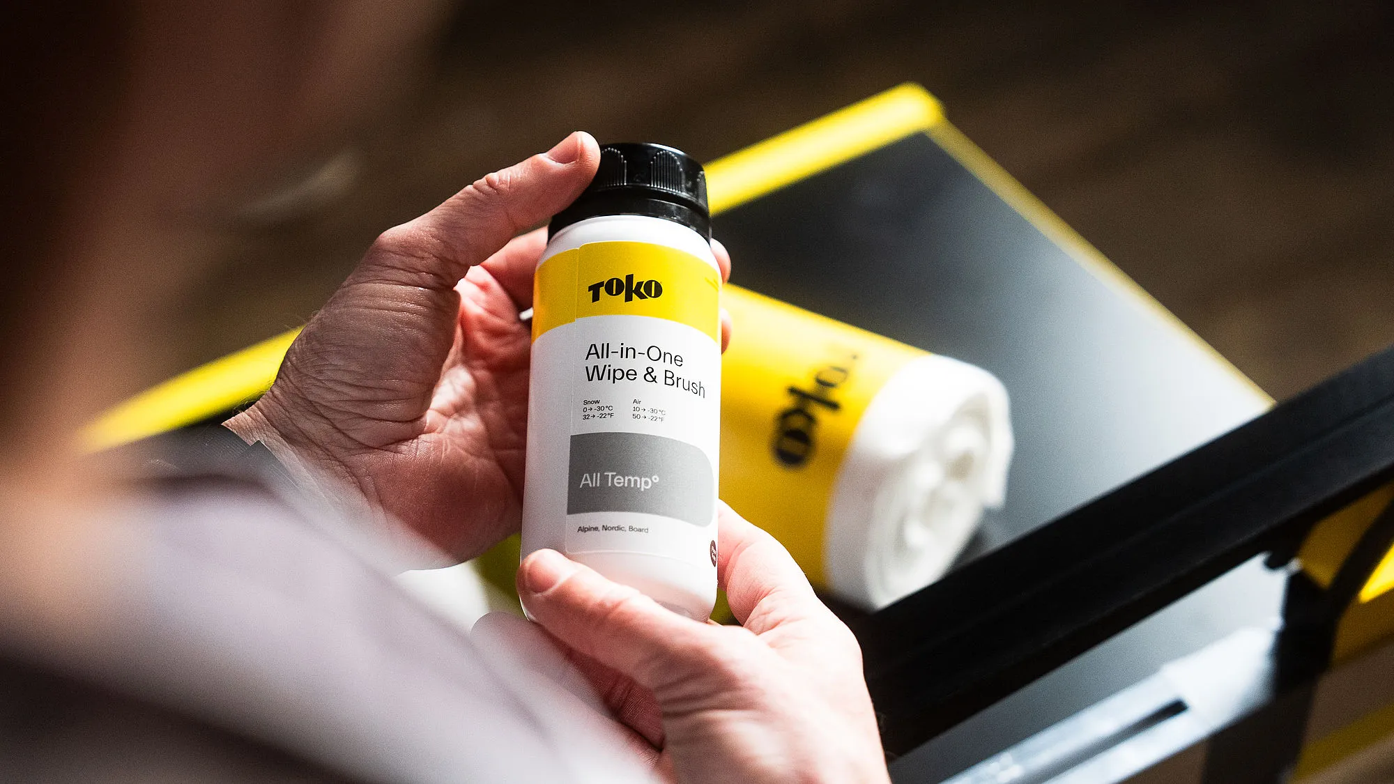 Une personne tient une bouteille "Toko All-in-One Wipe & Brush " devant un rouleau de lingettes nettoyantes. Les couleurs sont le jaune, le blanc et le noir.