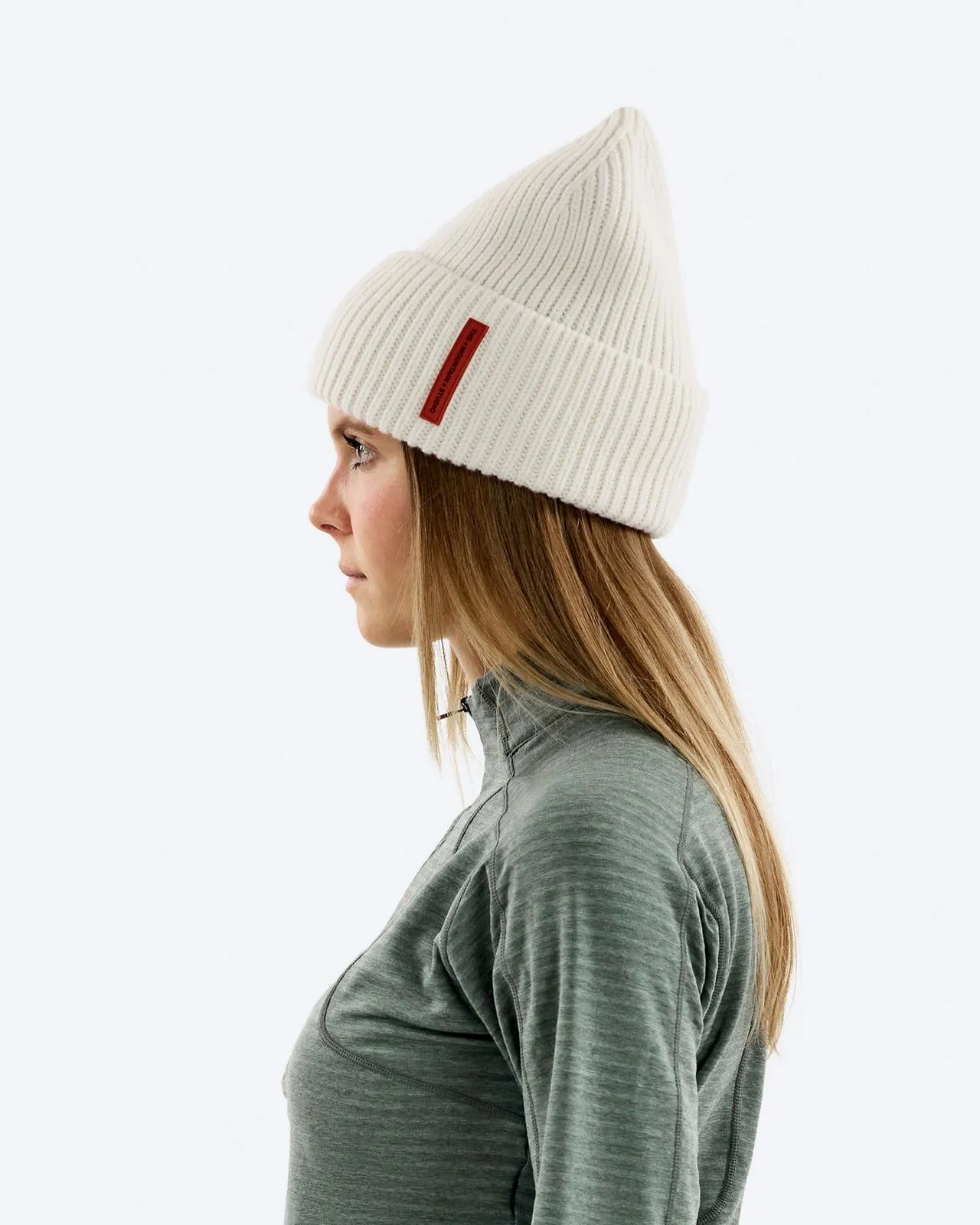 Profilo di una donna con capelli biondi che indossa un berretto bianco a coste con etichetta rossa e un top grigio.