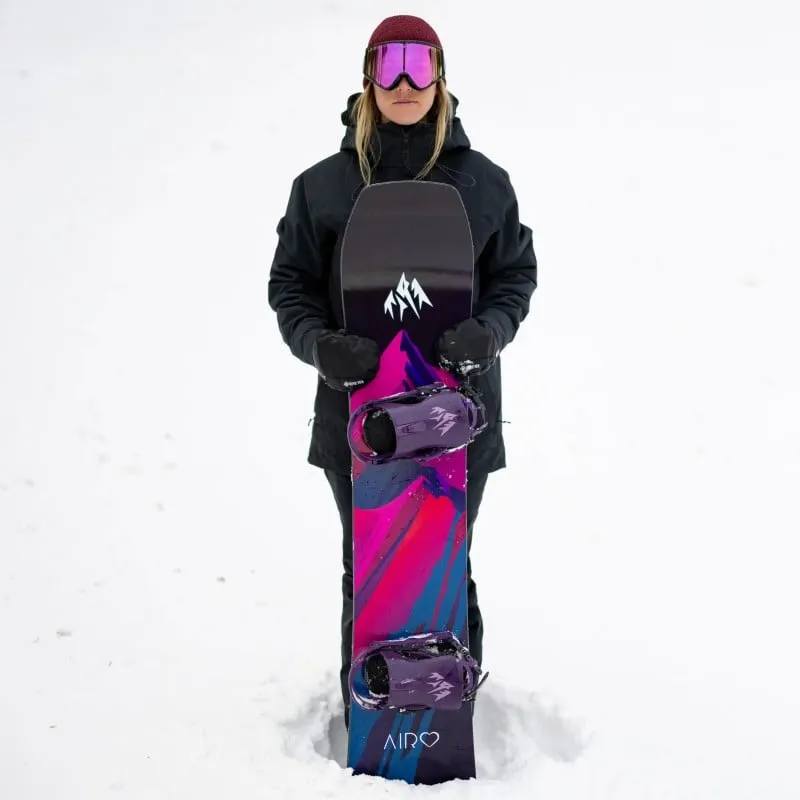 Femme avec masque de ski tient Jones Airheart 2.0 Snowboard avec un design coloré dans la neige.