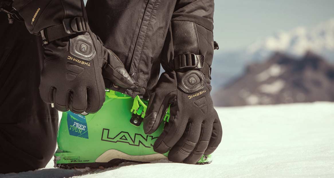 Personne agenouillée sur la neige, manipulant des chaussures de ski vertes avec des gants Thermic noirs.