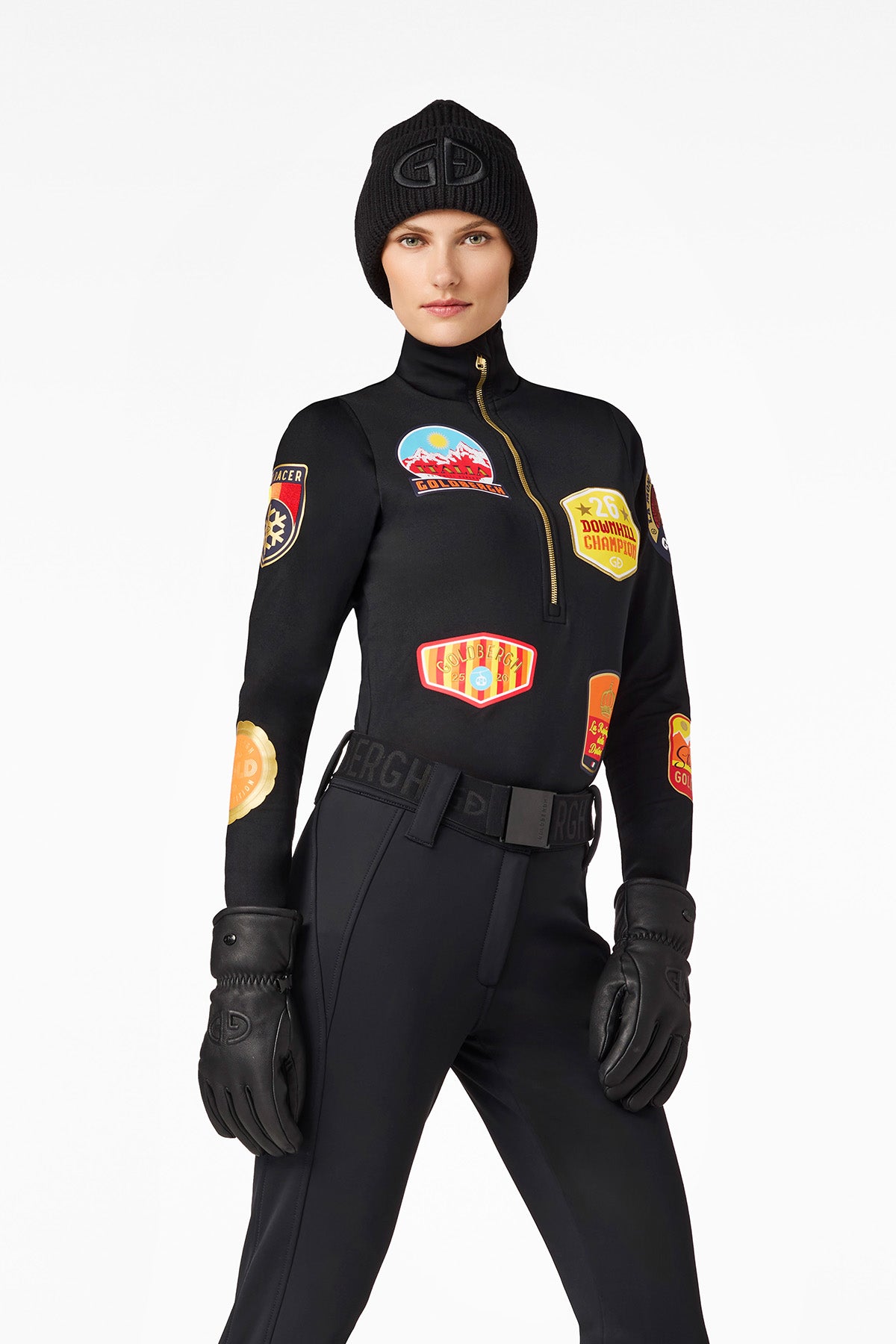 Woman in black ski clothing with patches, hat and gloves in front of a white background.