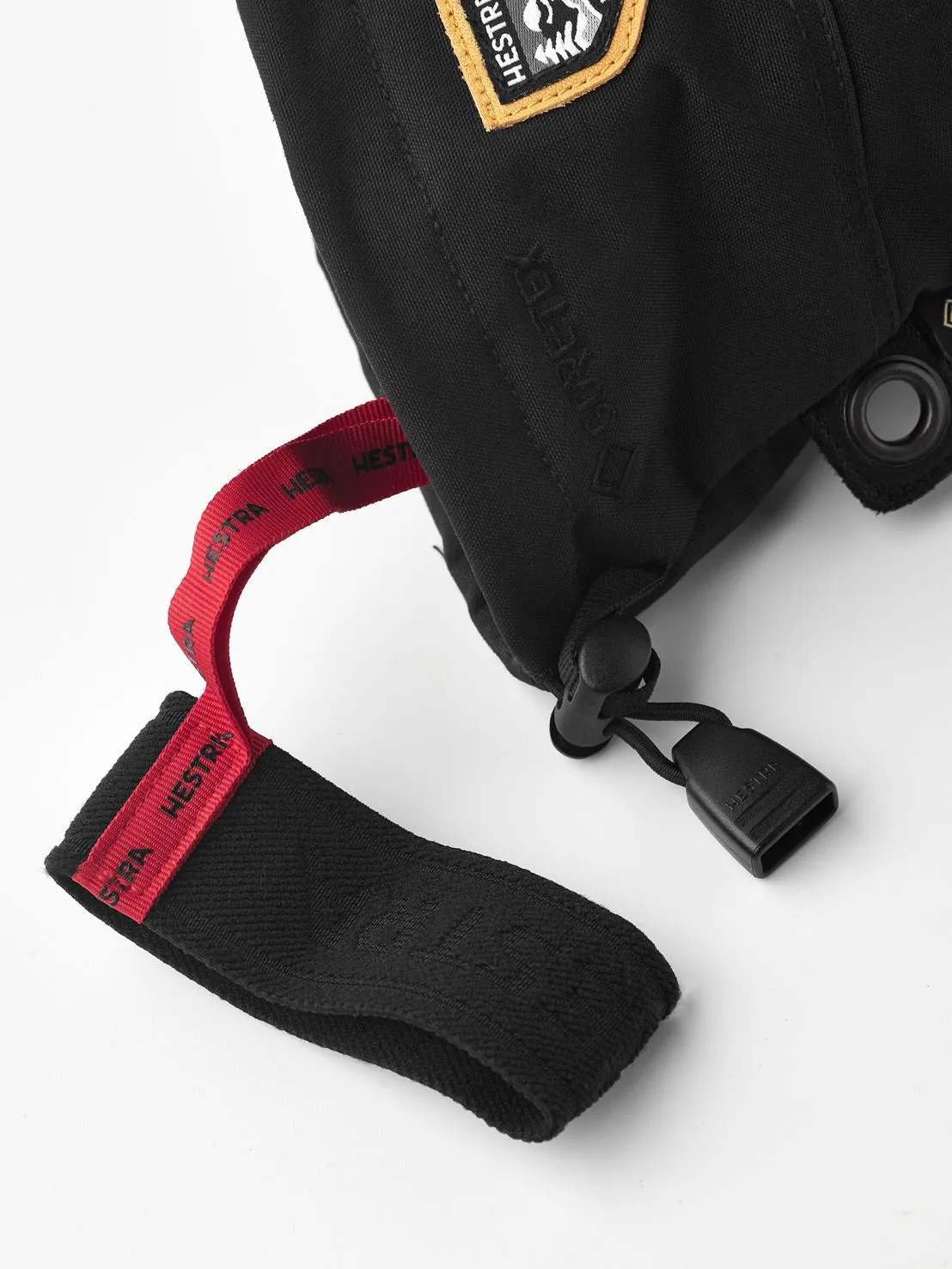 Close-up of a black Hestra mitten with Gore-Tex, red loop and logo patch. Details such as the drawstring and eyelet are visible.