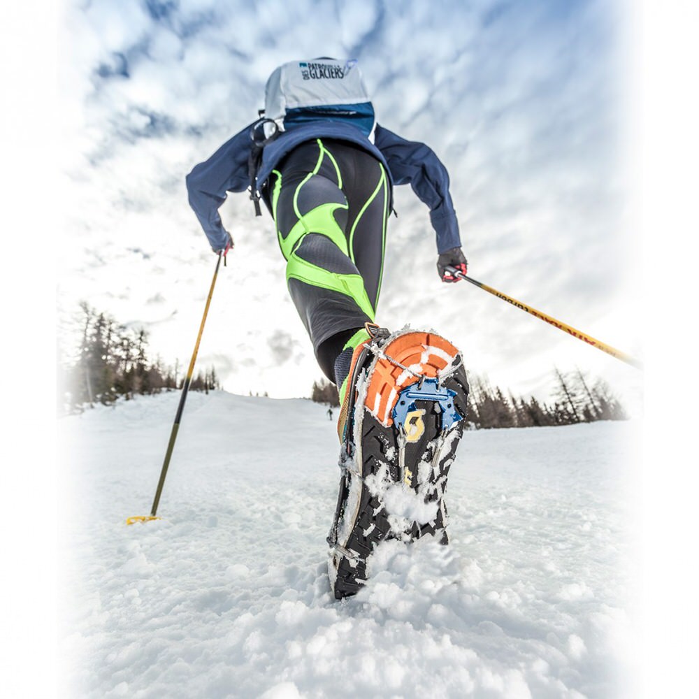 Une personne court en montée dans la neige avec une griffe de chaussure de trail, des bâtons et un sac à dos. La griffe permet de s'accrocher sur la glace et les surfaces glissantes.