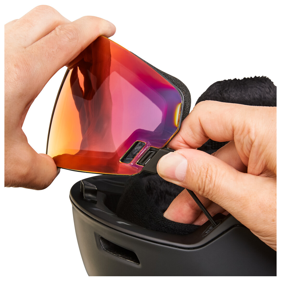 Eine Hand befestigt eine rote, getönte Skibrillenlinse an einem schwarzen Skihelm. Die Linse wird durch einen Clip an Ort und Stelle gehalten.
