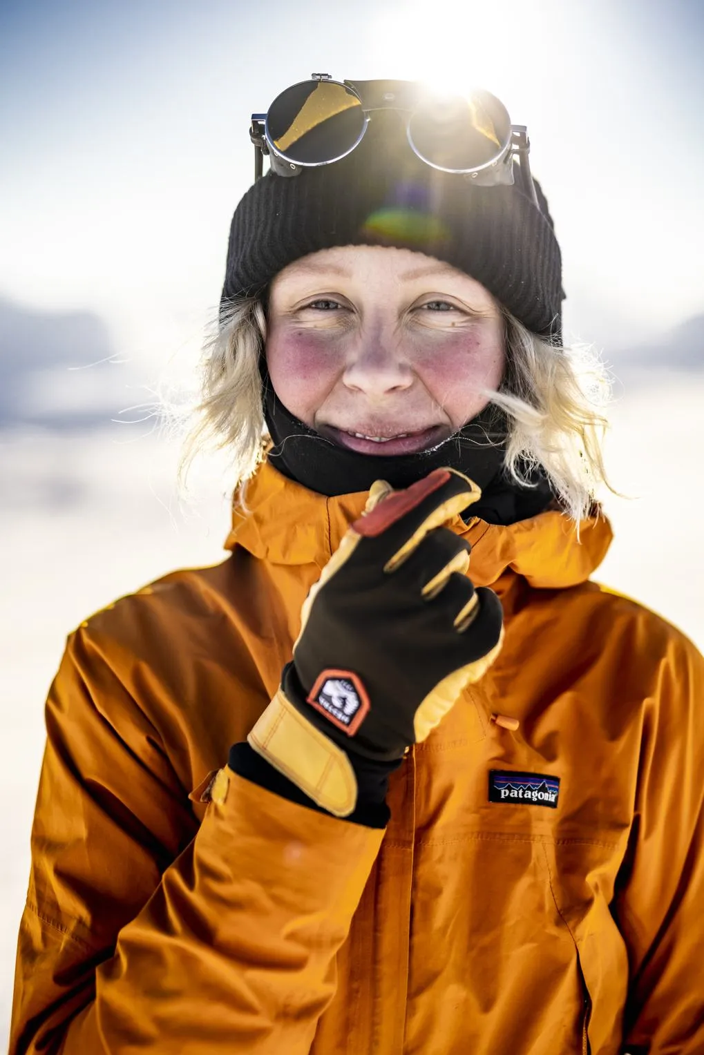 Personne souriante avec un bonnet et des lunettes de ski sur la tête, portant une veste orange et des gants dans la neige.