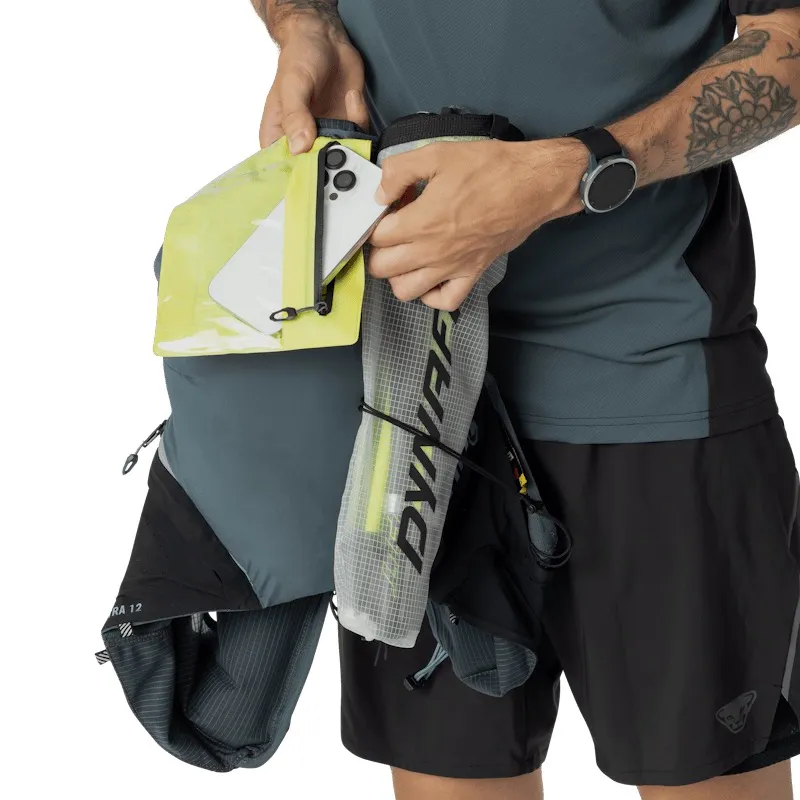 A man in athletic wear is placing a smartphone into the yellow, waterproof pocket of a running backpack. The backpack features compartments for poles and hydration. He is wearing a smartwatch on his wrist and has a tattoo on his upper arm.