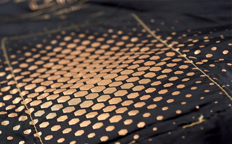 Detailaufnahme der Therm-ic Heated Vest Women. Muster aus goldenen Sechsecken auf dunklem Stoff, das in Punkte übergeht.