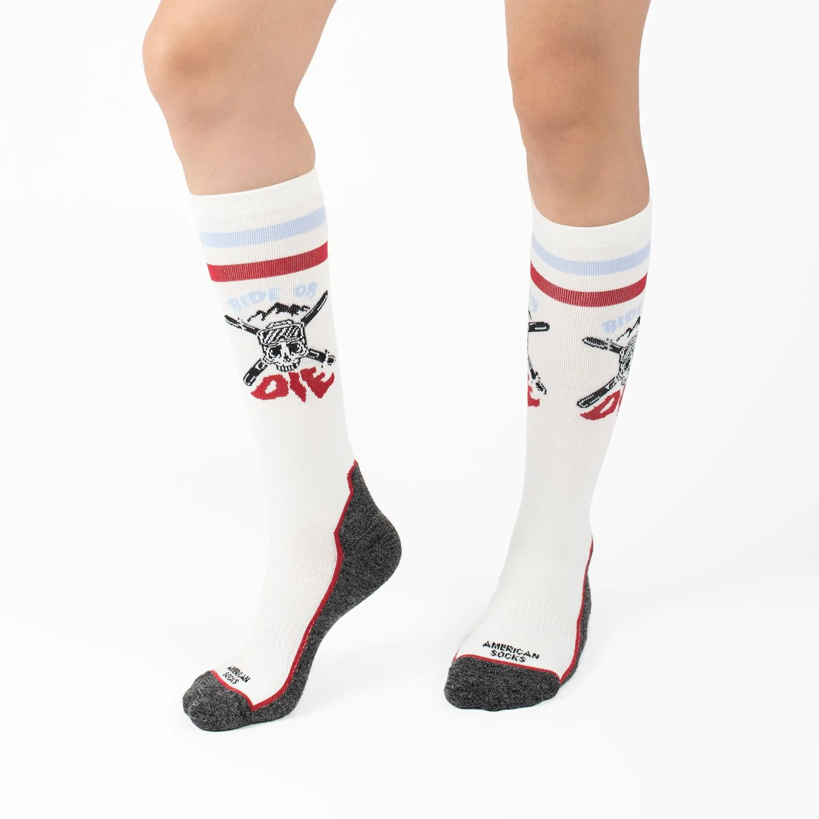 Gambe con calze da neve "Ride or Die Snow Socks" lunghe fino al ginocchio, bianche, con motivo a teschio, grigie sulle dita e sul tallone.