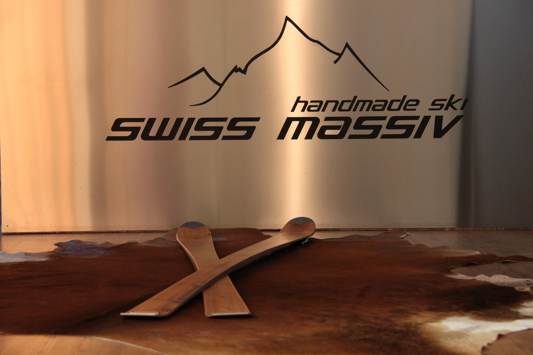 Swissmassiv Entlebucher Holz Skis | ski-shop.ch Zwei Holzski gekreuzt auf einem Fell, im Hintergrund ein Schild mit dem Schriftzug "SWISS MASSIV" und Bergmotiv.
