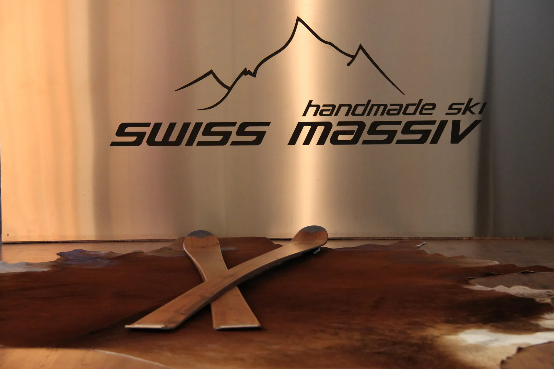 Due sci di legno incrociati su una pelle, sullo sfondo un'insegna con la scritta "Swiss Massiv" e un motivo di montagna.