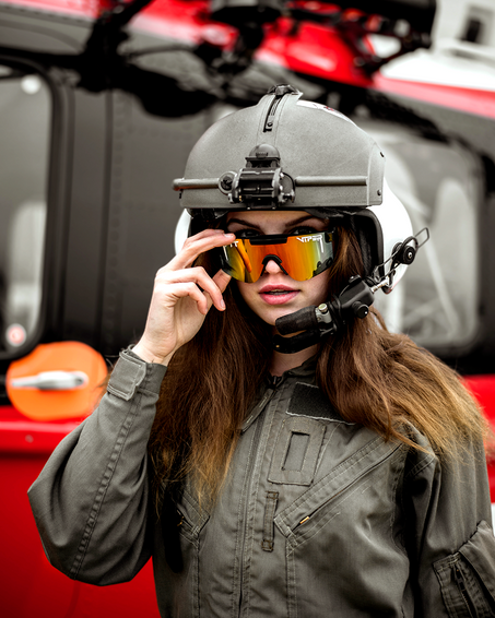 Frau vor Helikopter Frau mit Helm und polarisierter rosa-grauer Pit Viper Sonnenbrille vor rotem Helikopter.