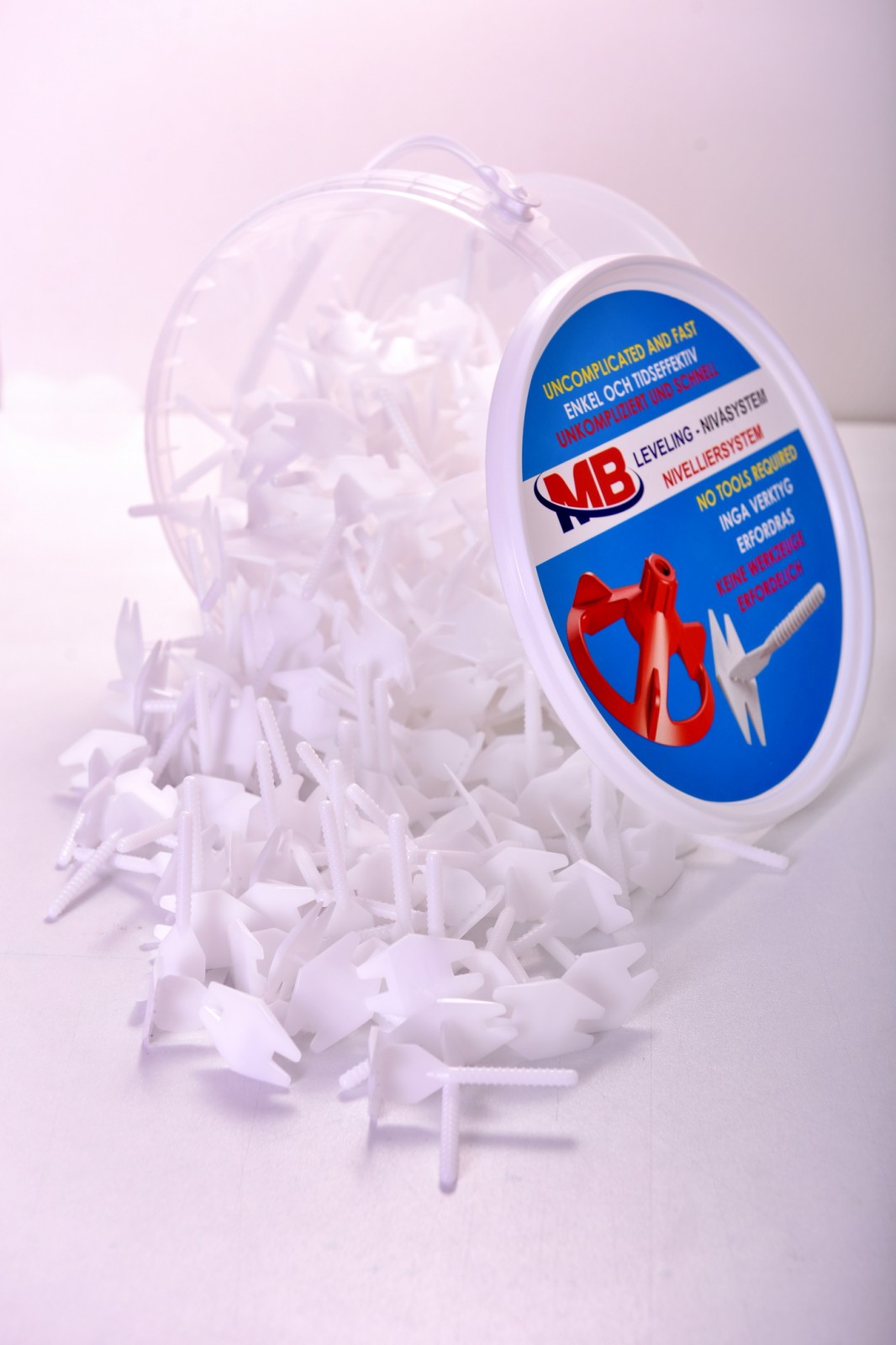 MB Leveling Pièces du système Des languettes filetées blanches du système MB Leveling se trouvent devant un récipient transparent avec une étiquette bleue et blanche.