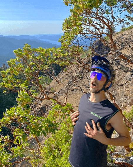 Man with helmet and pink glasses on a mountain, hands on his chest, green trees and a lake in the background. Afterparty 2000 feeling.